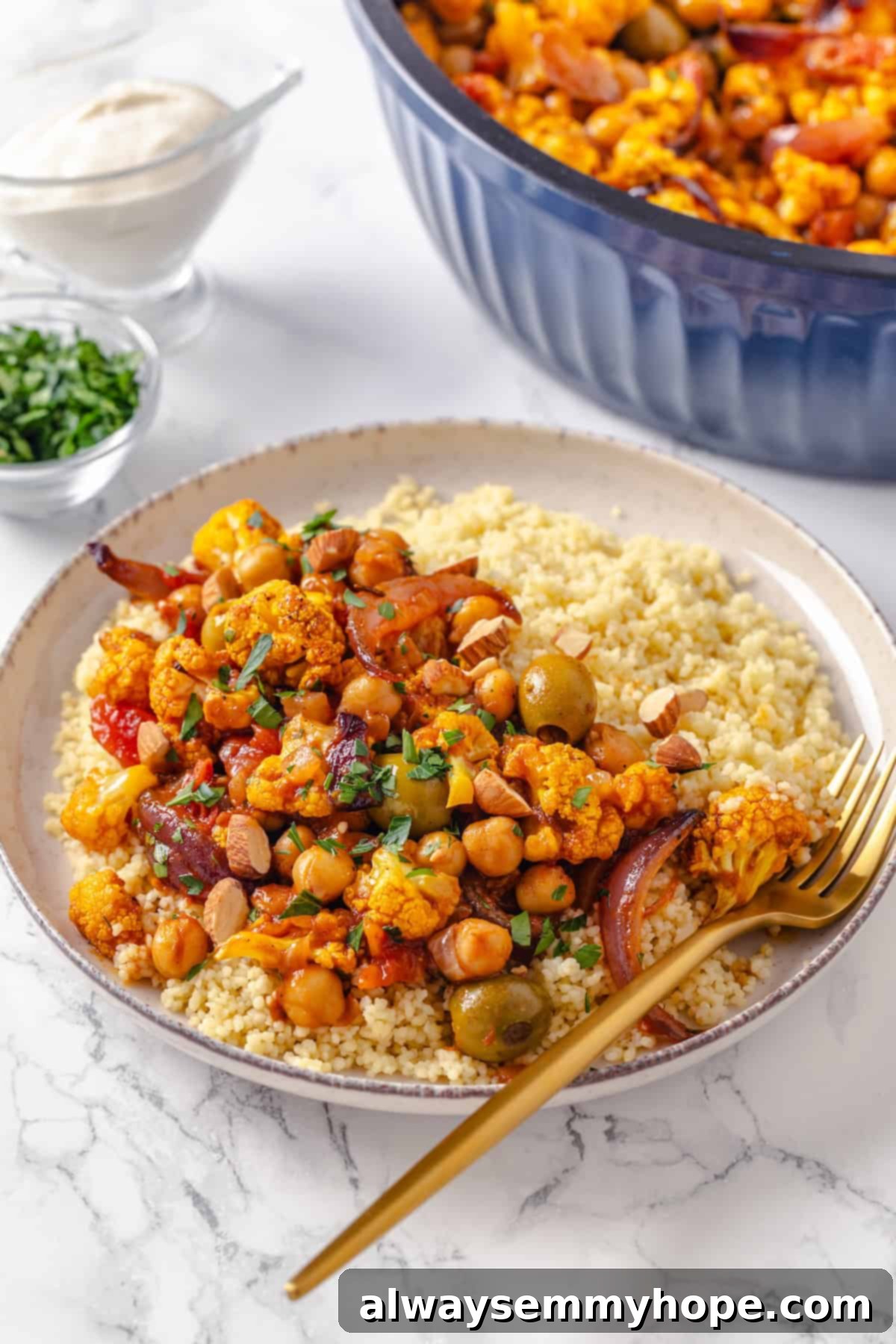 Warm spices mingle with vegetables, golden raisins, olives and chickpeas to make this Moroccan Cauliflower Tagine a delicious vegan dinner! Bowl of Moroccan cauliflower tagine with couscous and gold fork.