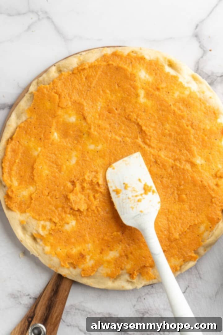 Spreading Creamy Sweet Potato Sauce onto the Pizza Crust Spreading creamy sweet potato sauce evenly onto a pre-baked pizza crust, creating a vibrant orange base.