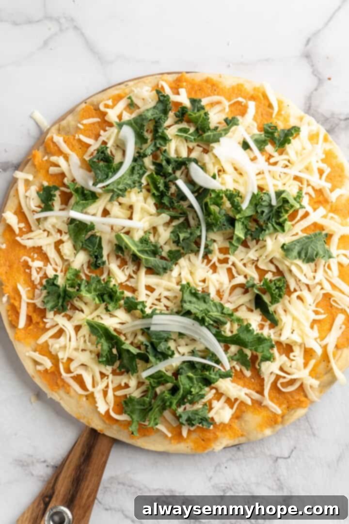 Topping and Baking the Sweet Potato and Kale Pizza Overhead view of an unbaked sweet potato and kale pizza, generously topped with fresh kale, thinly sliced onions, and shredded vegan mozzarella.