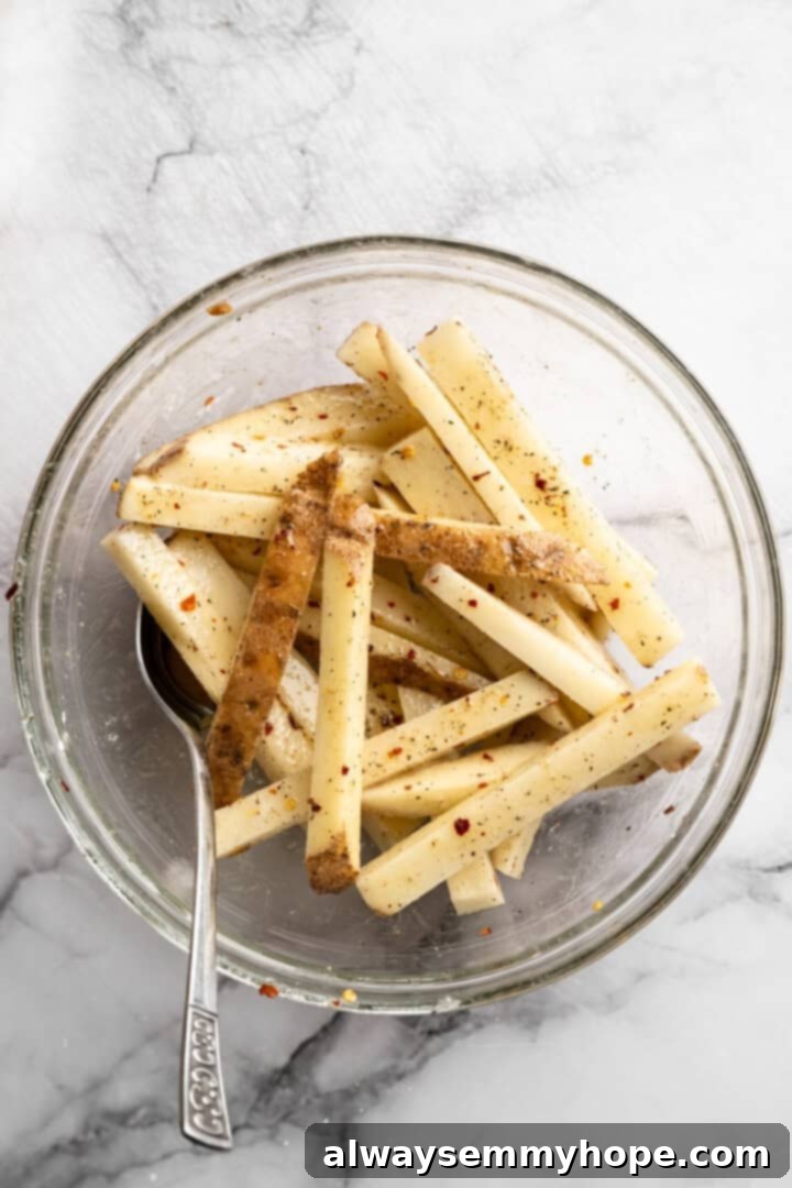 Seasoning the raw potato wedges with olive oil, cornstarch, and spices. Raw fries in glass bowl with spoon.