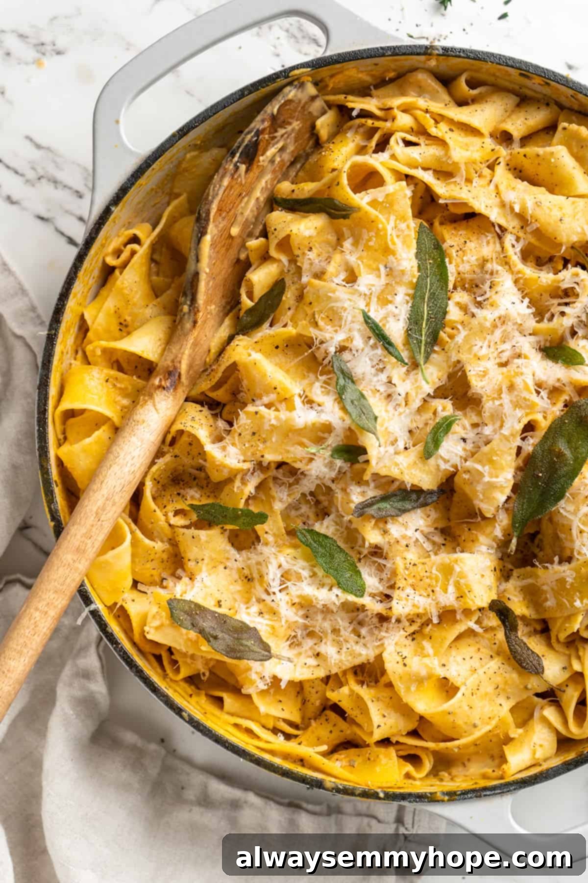 This easy pumpkin Alfredo pasta is rich, creamy, and perfect for a weeknight. Turn simple pantry ingredients into a restaurant-style meal! Overhead view of pumpkin alfredo pasta in skillet with wooden spoon and crispy sage leaf garnish