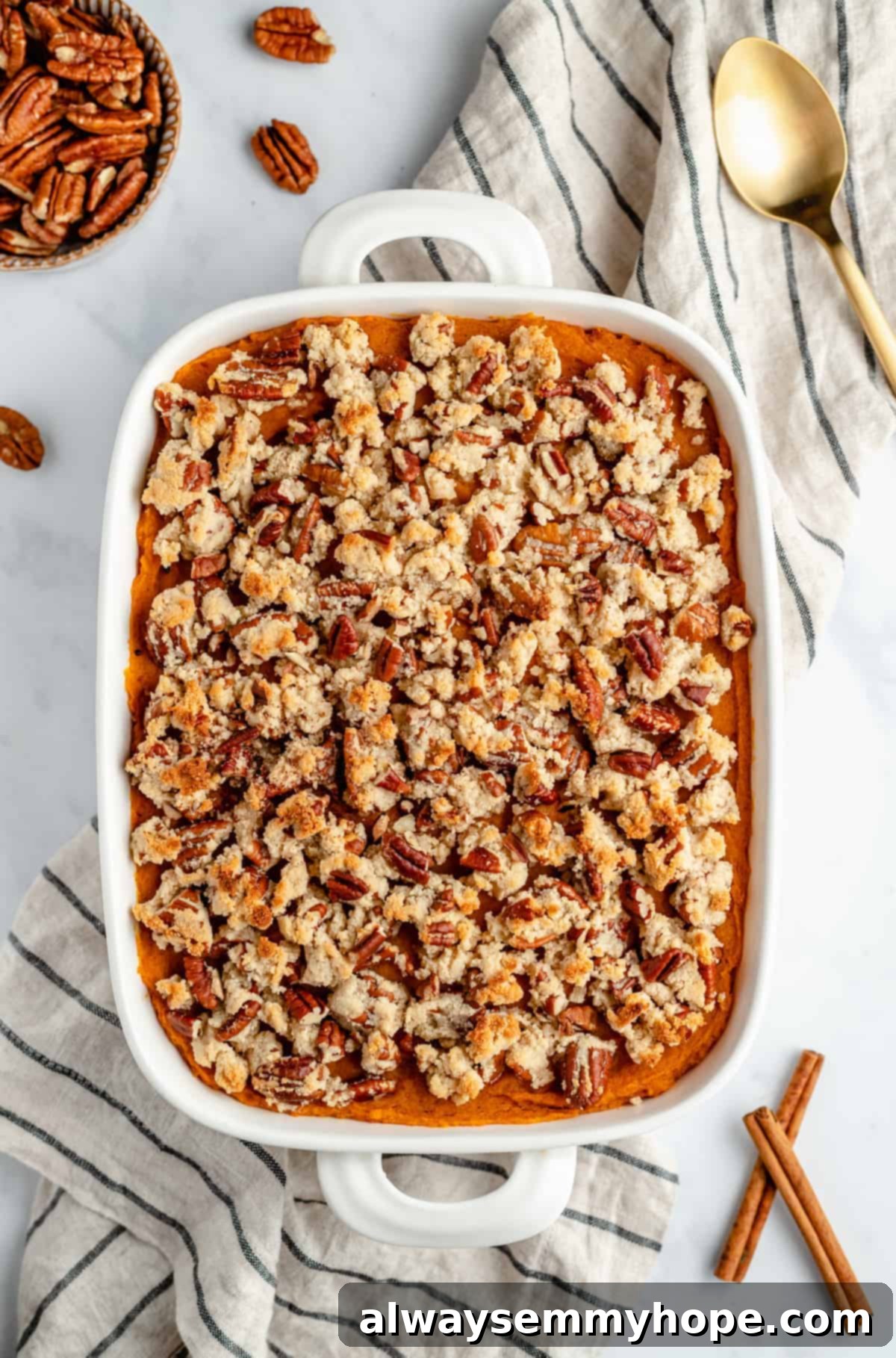 Maple Pecan Crunch Vegan Sweet Potato Bake 3 A cozy scene featuring a baking dish filled with Vegan Sweet Potato Casserole, adorned with a golden maple pecan topping, surrounded by fresh ingredients like whole pecans and a cinnamon stick on a striped kitchen towel.