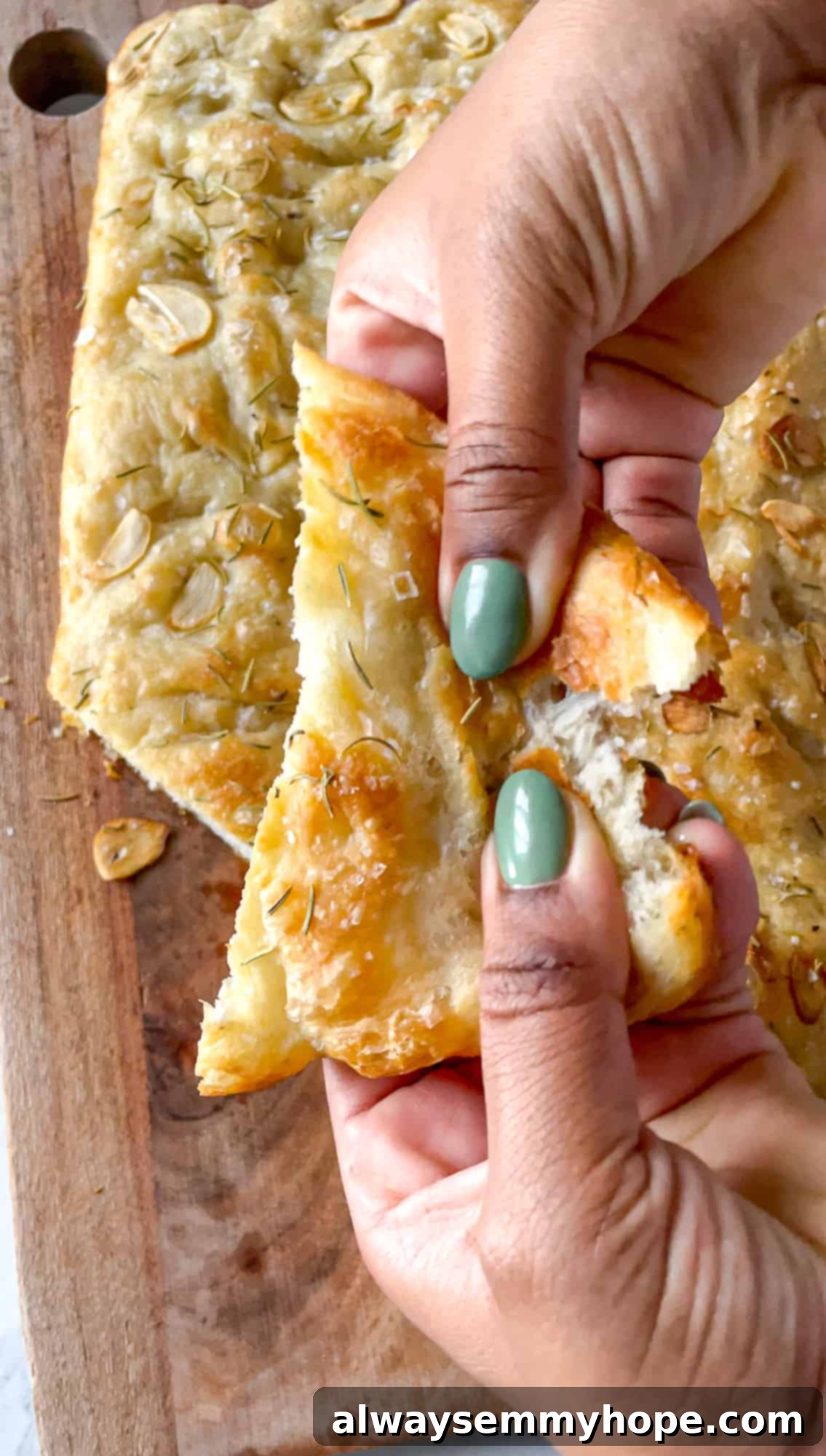 Two hands gently tearing a slice of freshly baked garlic and rosemary focaccia bread, revealing its soft interior and crispy crust.