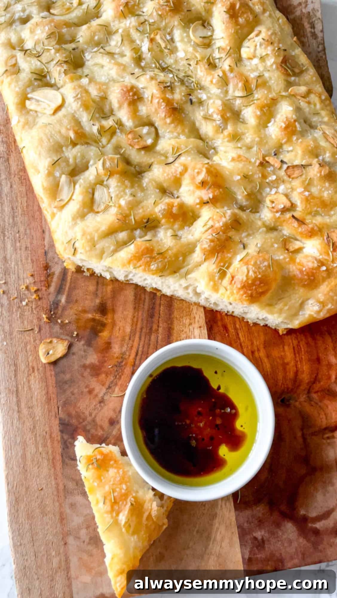 A large slice of garlic and rosemary focaccia bread, freshly cut and arranged on a wooden cutting board, with a small bowl of olive oil and balsamic vinegar for dipping beside it.