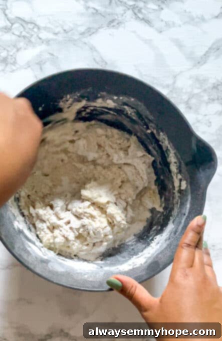 A person's hands stirring flour, yeast, sugar, and salt in a large mixing bowl to combine them thoroughly.