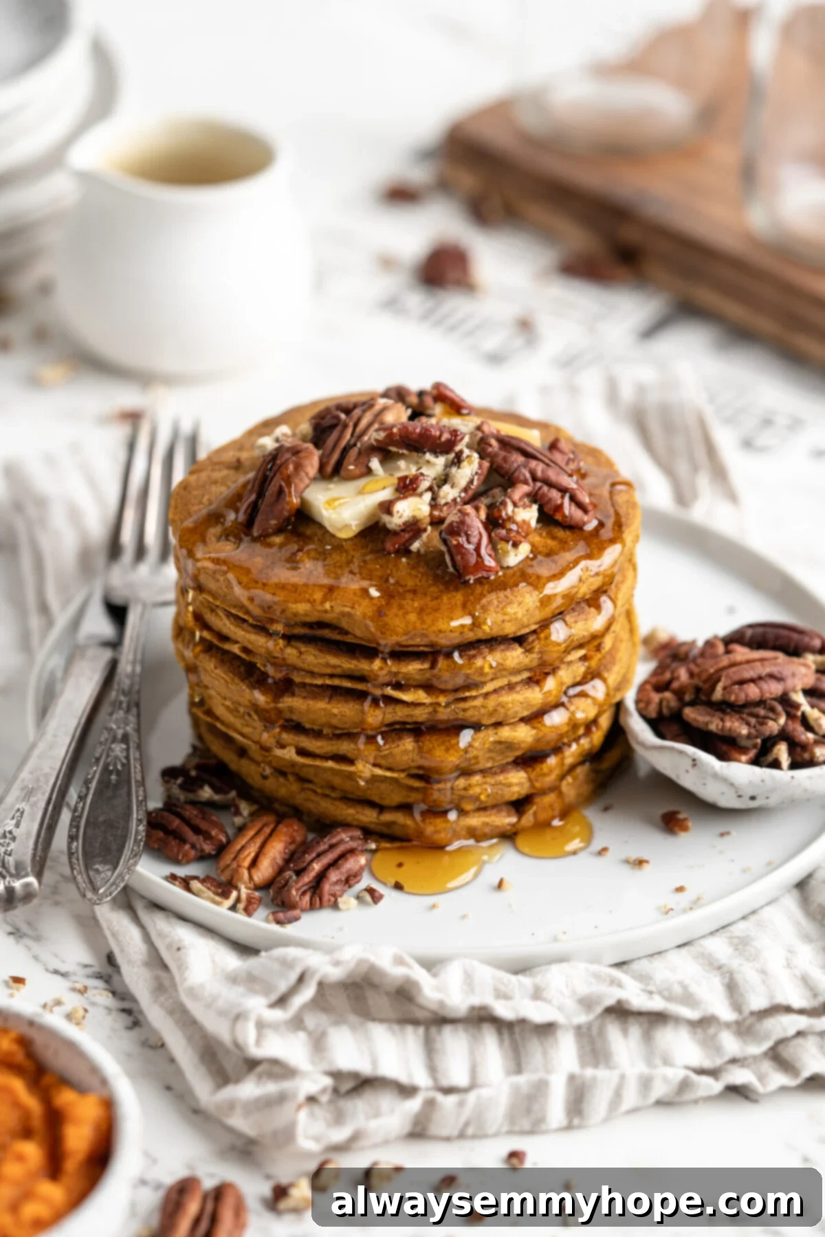 A beautifully stacked plate of fluffy vegan gluten-free pumpkin pancakes, generously topped with pecans and a drizzle of maple syrup. A small bowl of extra nuts and a fork are beside the plate, all ready for a delicious fall breakfast.