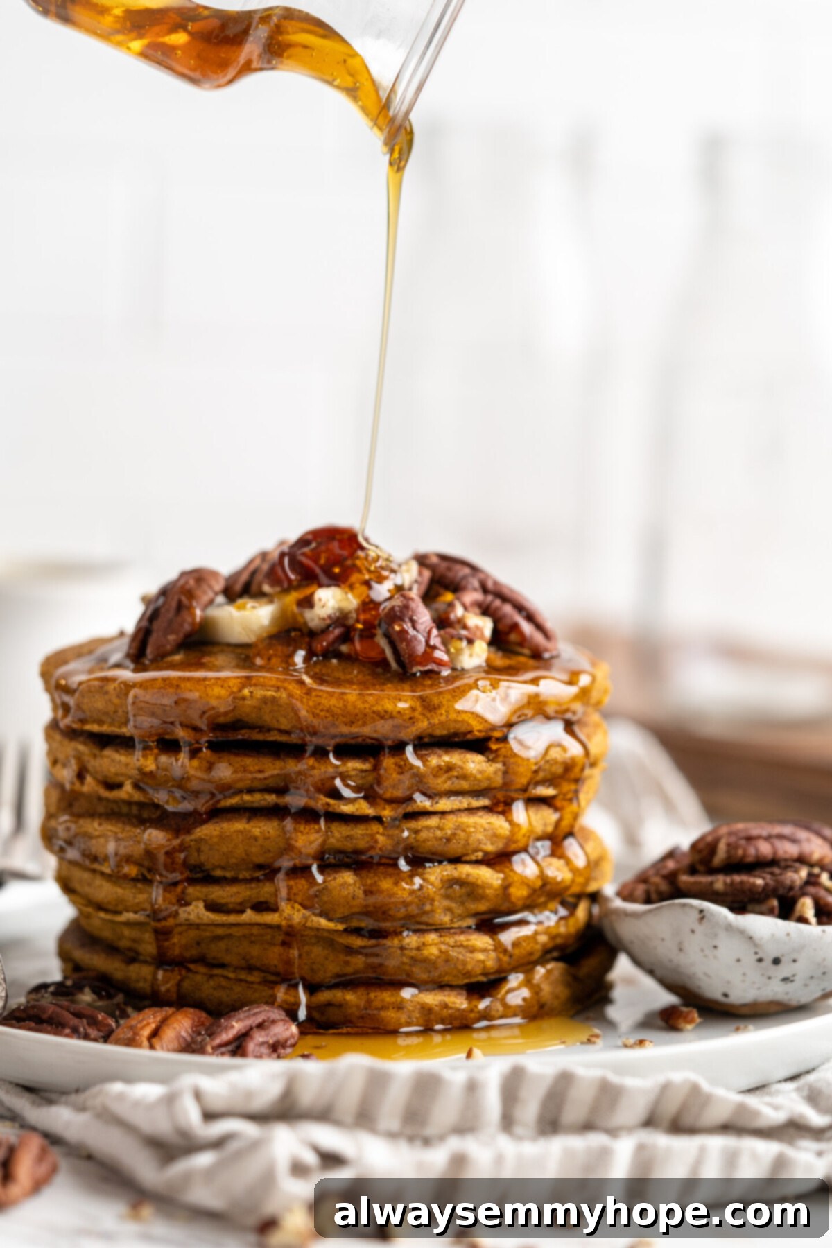 A delectable stack of golden-brown vegan gluten-free pumpkin pancakes, generously drizzled with warm maple syrup and topped with a scattering of chopped nuts. The syrup is captured mid-pour, adding a dynamic touch.
