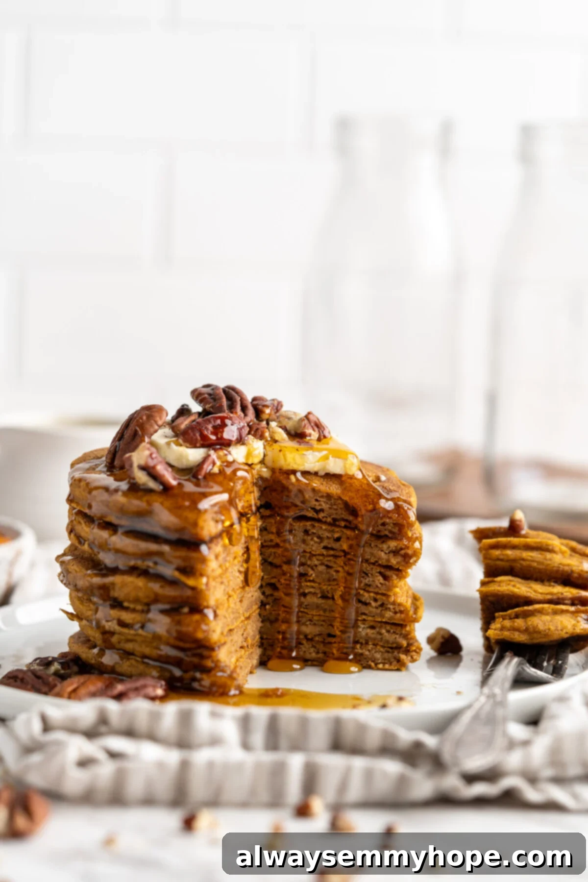 A delicious stack of vegan gluten-free pumpkin pancakes on a white plate, beautifully topped with a variety of nuts. One quarter of the stack has been cut out, revealing the fluffy, moist interior.