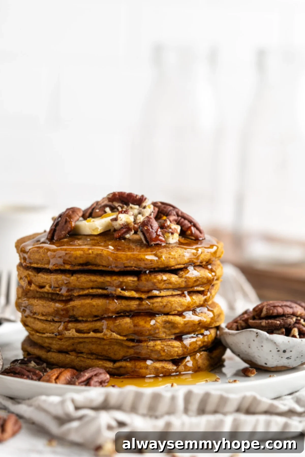 A close-up view of a tall stack of golden-brown vegan gluten-free pumpkin pancakes, garnished with a sprinkle of chopped nuts and ready to be drizzled with syrup. A small bowl of nuts sits nearby.
