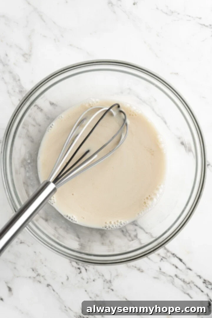 Overhead shot of a mixing bowl containing a clear liquid, likely almond milk, with a whisk resting in it. This showcases the initial step of combining wet ingredients.