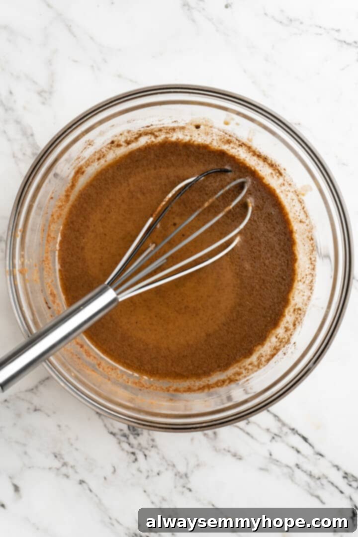 Overhead view of a mixing bowl with vibrant orange pumpkin puree, brown cinnamon, and white almond milk being whisked together, illustrating the blending of wet ingredients and spices for pumpkin pancakes.