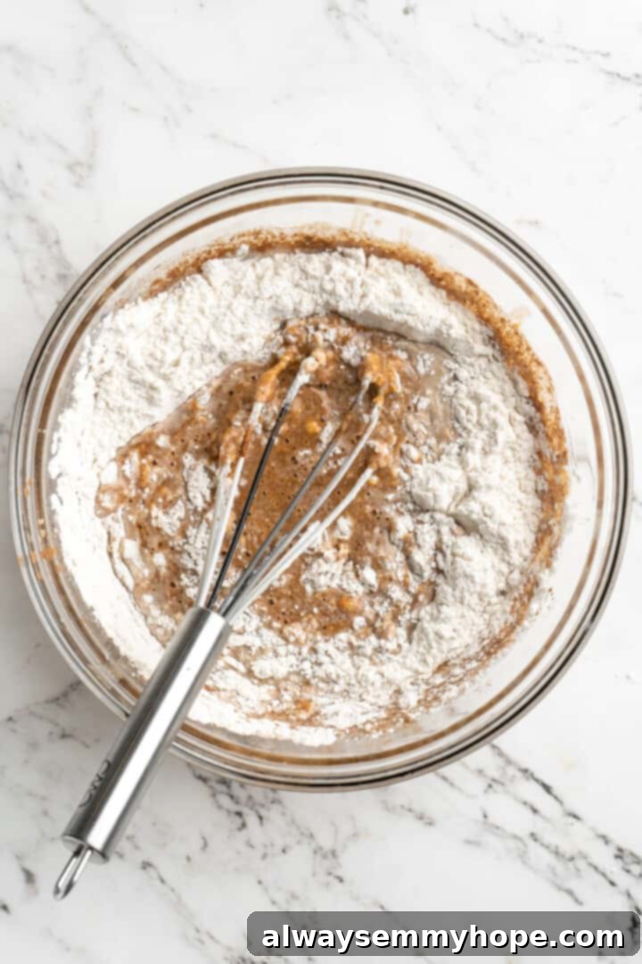 Overhead shot of a whisk mixing light flour into the vibrant orange pumpkin puree and almond milk mixture in a bowl, demonstrating the addition of dry ingredients to the wet.