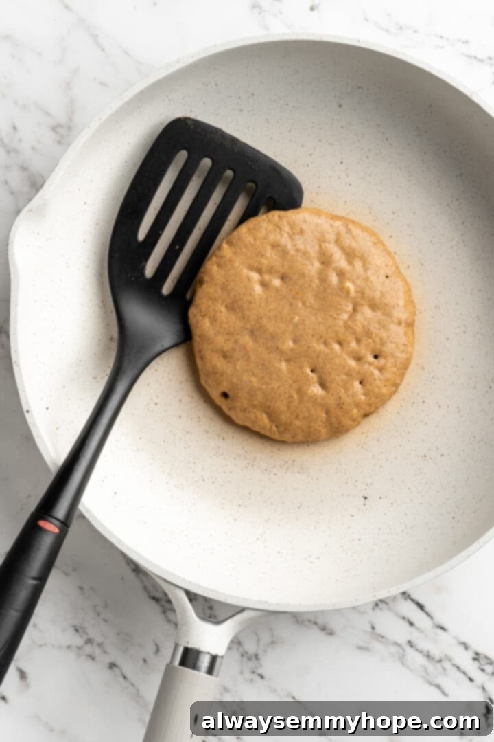 Overhead view of a perfectly round, uncooked vegan pumpkin pancake sizzling in a pan, with a spatula poised to flip it. Bubbles are visible on the surface.