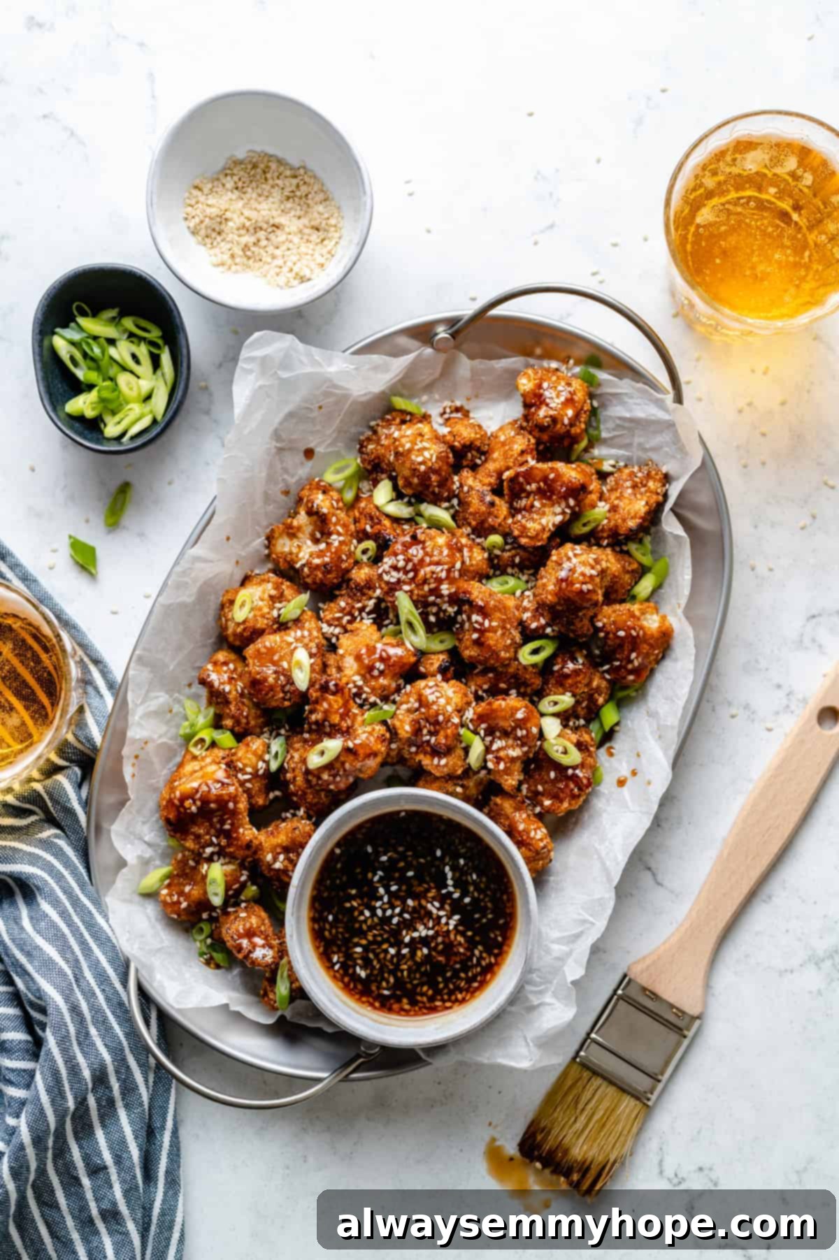 plate of cauliflower wings in a tray with sauce in a small bowl beside it surrounding by toppings and a brush to brush sauce on wings
