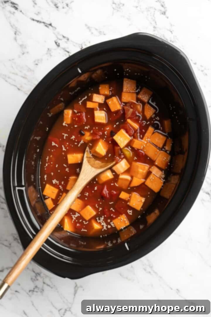 Overhead view of sweet potatoes, black beans, tomatoes, and broth in slow cooker insert with wooden spoon