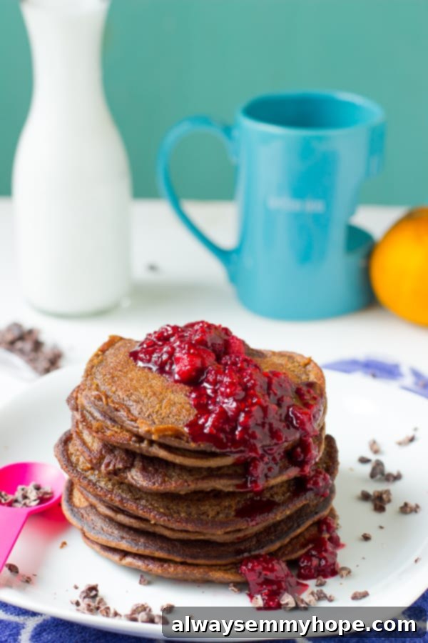A stack of fluffy chocolate peanut butter pancakes, drizzled with bright red raspberry compote, on a pristine white plate, ready to be enjoyed.