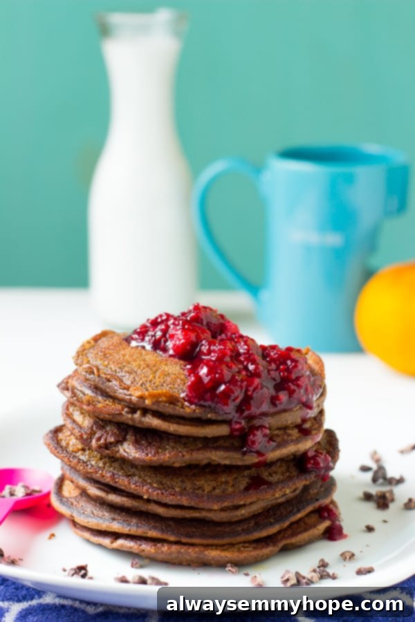 A side shot of a tall stack of chocolate peanut butter pancakes, richly coated with raspberry compote and fresh berries, presented on a white plate.