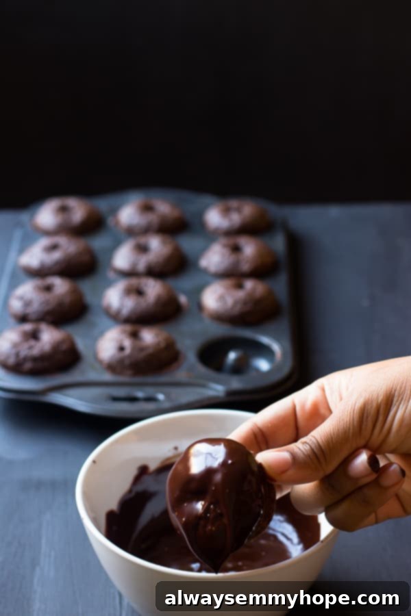 A hand carefully dipping a baked chocolate s'mores donut into a rich, glossy chocolate glaze.