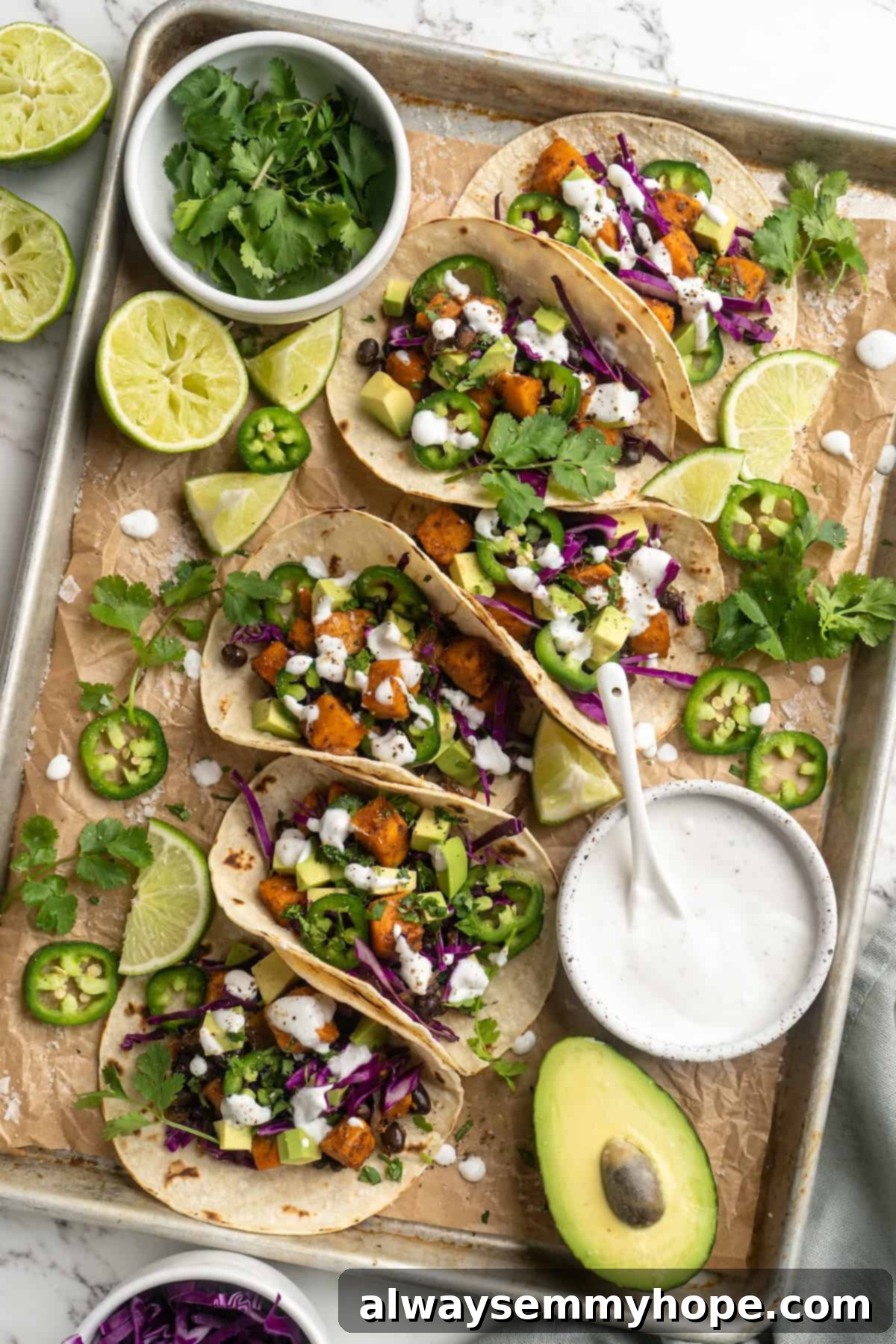 Overhead view of several assembled sweet potato and black bean tacos on a parchment-lined baking sheet, surrounded by fresh toppings like avocado, cilantro, and lime, ready for serving