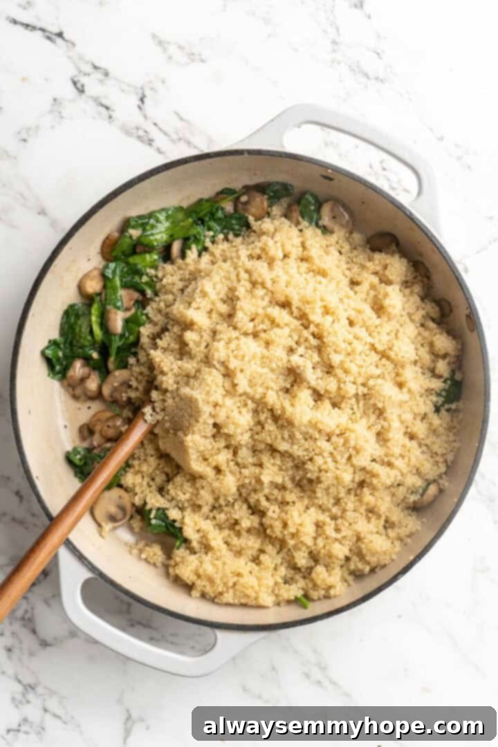 Overhead view of cooked quinoa being stirred into the skillet with sautéed spinach and mushrooms, ready to be combined.