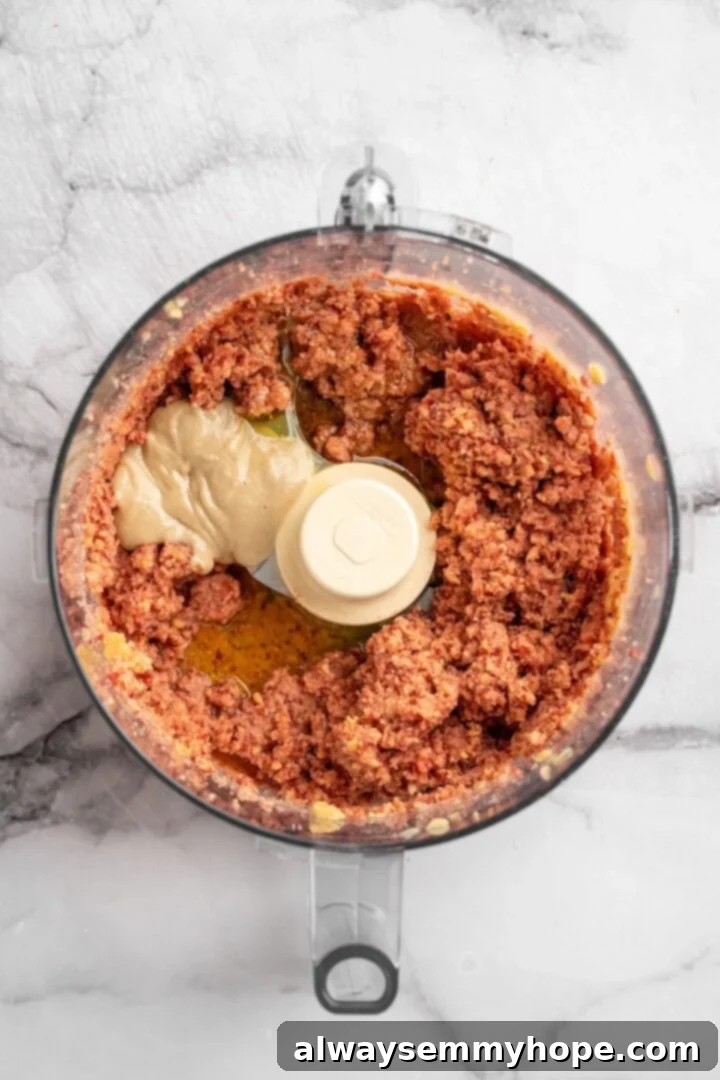Step 2: Add tahini, olive oil, lime juice, garlic, and seasonings to the food processor for the beet hummus. Tahini being added to the food processor bowl containing pulsed beets and chickpeas, alongside other ingredients.