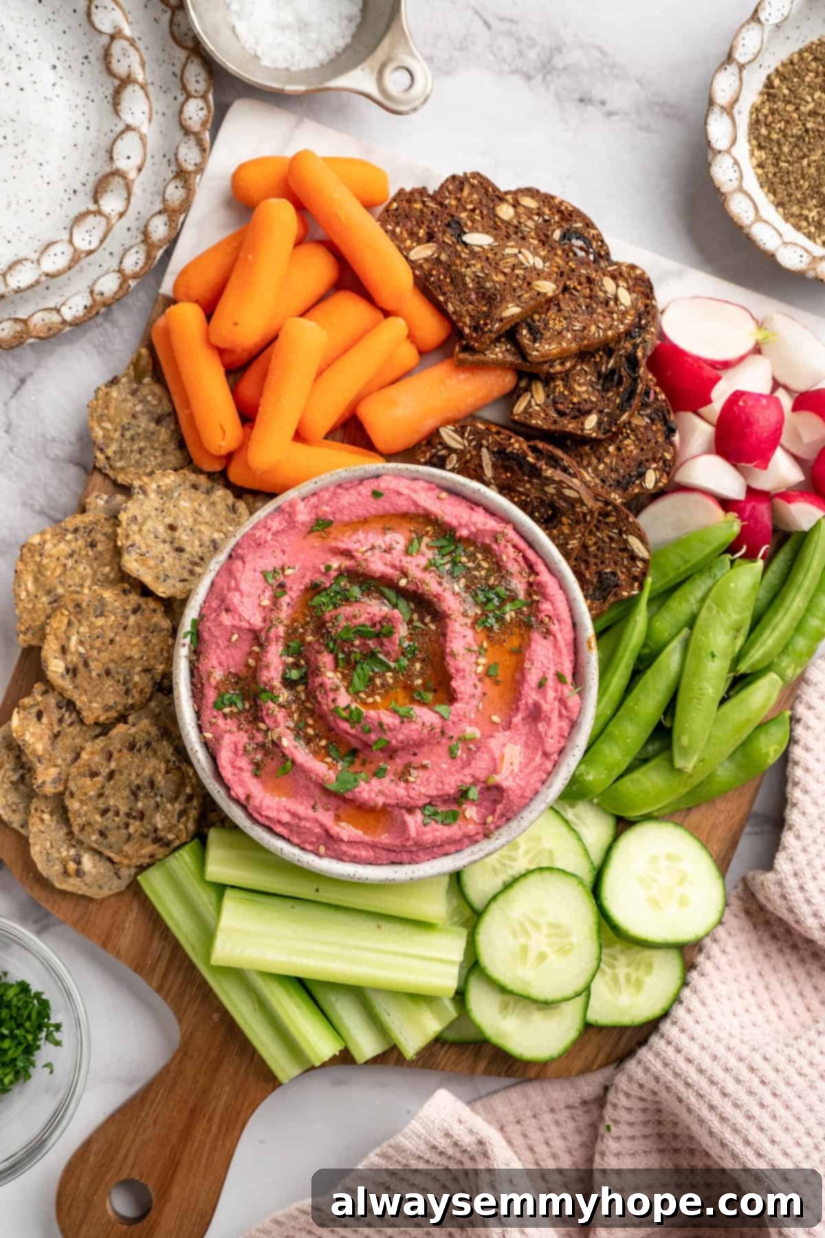 A beautifully arranged snack board showcasing creamy beet hummus served with an assortment of dippers. Wooden board featuring a bowl of vibrant beet hummus, surrounded by assorted crackers and fresh raw vegetables like carrots, cucumbers, and bell peppers.