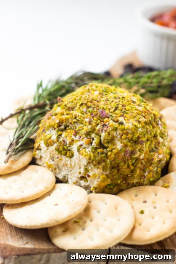 Pistachio Crowned Vegan Cheese Ball 4 Side on shot of pistachio crusted vegan cheese ball with a sprig of rosemary and crackers on the side.