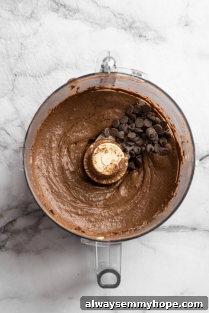 This Chocolate Avocado Pudding is ultra rich and decadent, with the creamiest texture and deepest chocolate flavour. Just 10 minutes of prep! A close-up of the smooth chocolate avocado pudding in the food processor bowl, with whole chocolate chips visible on the surface, ready to be pulsed in.