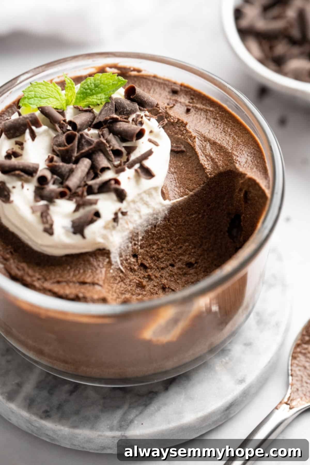 This Chocolate Avocado Pudding is ultra rich and decadent, with the creamiest texture and deepest chocolate flavour. Just 10 minutes of prep! A glass bowl of rich chocolate avocado pudding, generously topped with fluffy coconut whipped cream and delicately grated chocolate shavings, ready to be enjoyed.