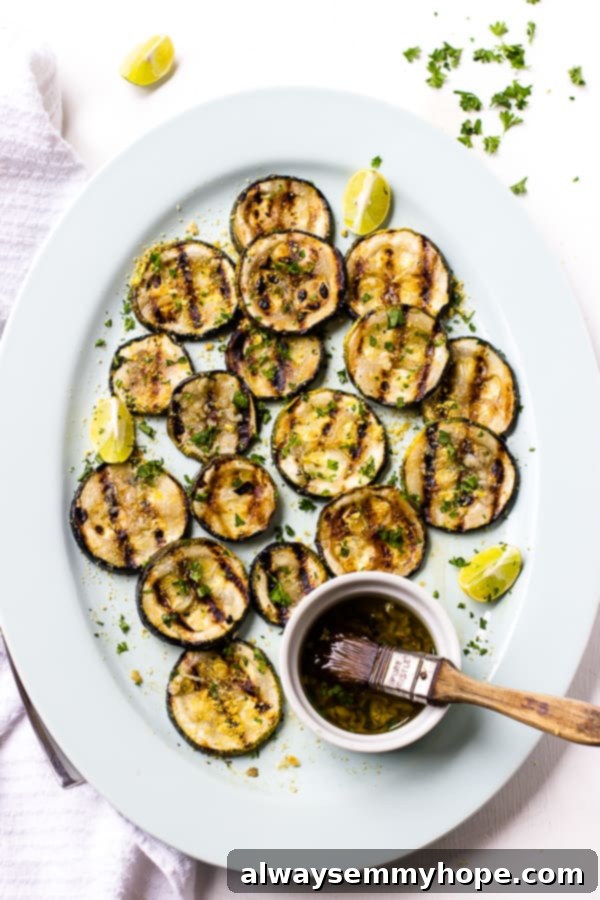 Overhead view of perfectly grilled lemon garlic zucchini slices on a serving platter with a small bowl of marinade and a basting brush nearby.