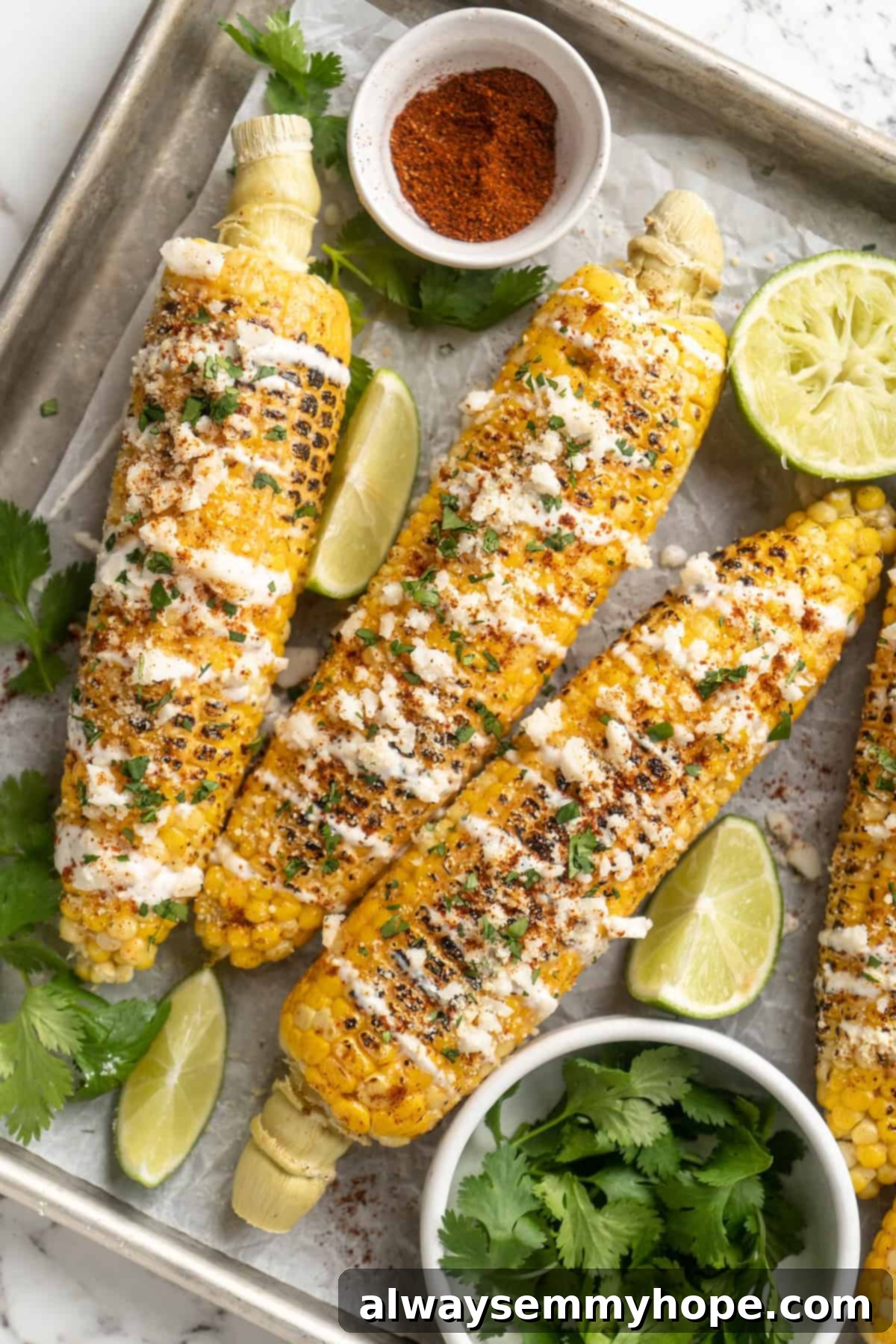 Delicious Vegan Elotes: Grilled Mexican Street Corn with Creamy Lime Crema. Overhead view of Vegan Elotes on a sheet pan with fresh cilantro, chili powder, and lime wedges as garnishes.