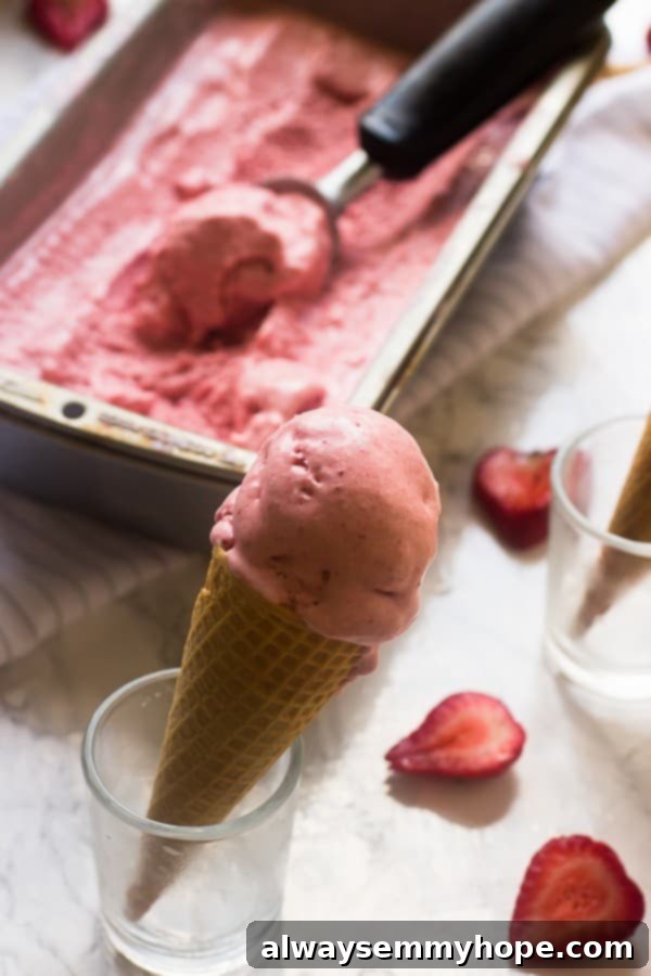Vegan strawberry coconut ice cream served in a waffle cone, with sliced fresh strawberries blurred in the background.