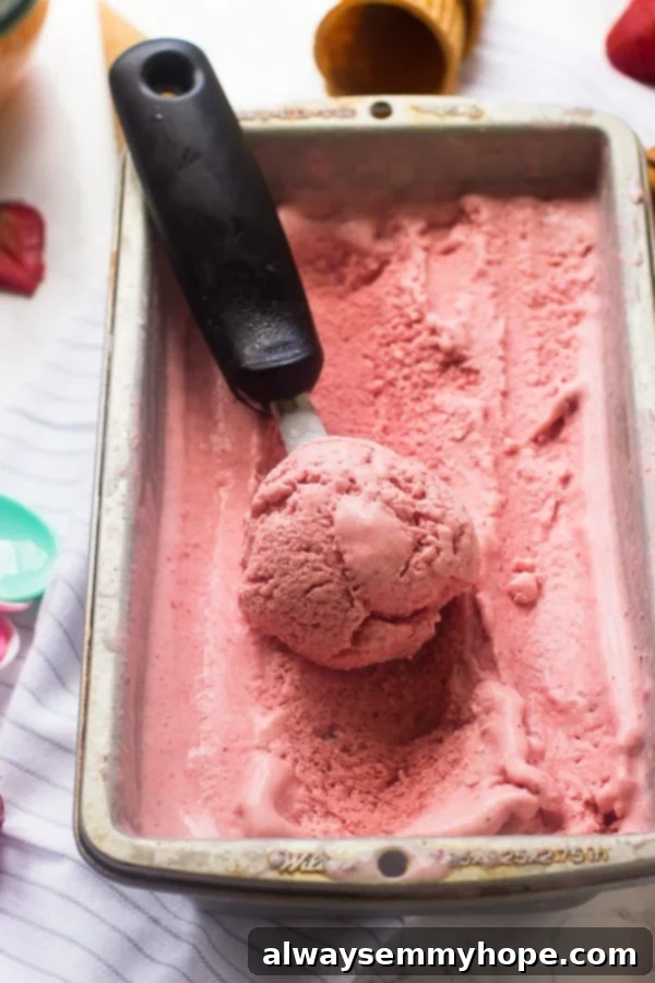 A vibrant tub of strawberry coconut ice cream with a silver scooper, showcasing its rich, creamy texture.
