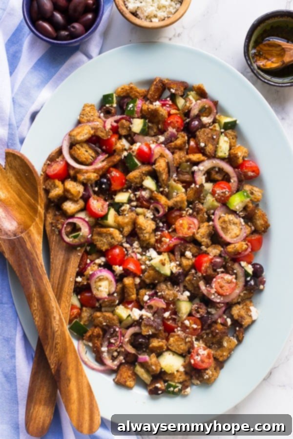 This super easy, gluten-free, vegan Greek panzanella salad is full of olives, cucumbers, and toasted bread, with a balsamic dressing. Top down shot of easy Greek panzanella salad on a white plate with wooden spoons on the side, showcasing toasted bread, cherry tomatoes, cucumbers, olives, and vegan feta.