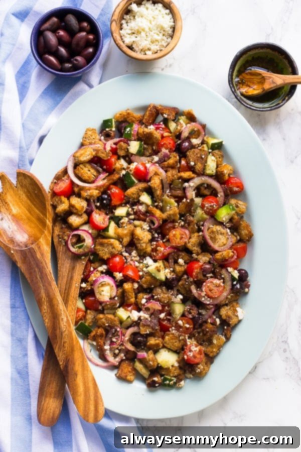 This super easy, gluten-free, vegan Greek panzanella salad is full of olives, cucumbers, and toasted bread, with a balsamic dressing. Top down shot of Greek panzanella salad on a white dish, with small bowls of olives, olive oil, and crumbled vegan feta cheese arranged neatly on the side.