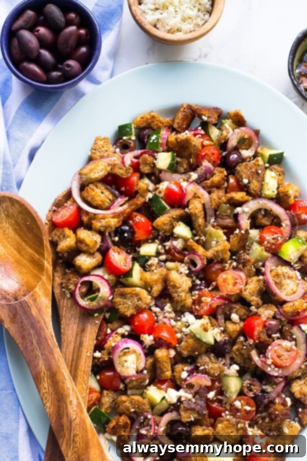 This super easy, gluten-free, vegan Greek panzanella salad is full of olives, cucumbers, and toasted bread, with a balsamic dressing. Top down shot of Greek panzanella salad in a white bowl with wooden spoons on the side, highlighting the fresh ingredients and toasted bread.